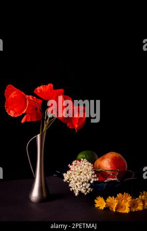 Stillleben auf schwarzem Hintergrund. Ein Strauß wilder roter Mohnblumen in einem Leuchtfeuer. Schale mit Früchten - Granatäpfel, Apfel und Avocado. Speicherplatz kopieren. Stockfoto