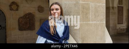 Junge Frau mit Schal auf blauem Trenchcoat, die in der Nähe des historischen Gebäudes in Wien posiert, Banner Stockfoto