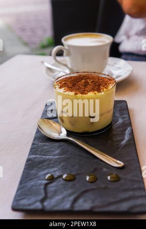 Tiramisu süßes Dessert in Italien mit einem Löffel auf einem Tisch mit einem defokussierten Hintergrund einer Tasse heißen Cappuccino Kaffee serviert. Stockfoto
