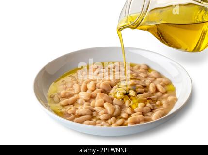 Extra natives Olivenöl, das aus einer Glaskanne über eine Bohnensuppe in einer weißen Schüssel abgefüllt wird, isoliert auf weiß, mit Schnittpfad im Preis inbegriffen Stockfoto