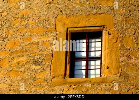 Alte gelbe Steinaußenwand und braune Holzfenster mit weißen Vorhängen. Absplitternde Stuckoberfläche. Verschlissenes Mauerwerk. Sandig-grobkörnige Textur. Stockfoto