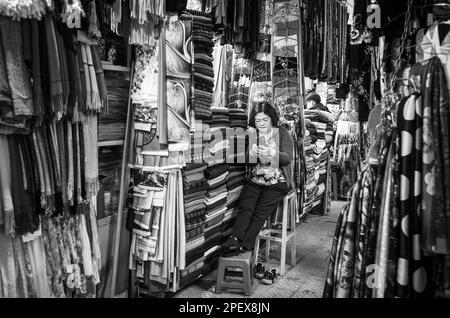 Eine vietnamesische Stallhalterin schaut auf ihr Mobiltelefon, während sie an ihrem Stoffstand im Dalat Market, Vietnam, auf Kunden wartet. Stockfoto