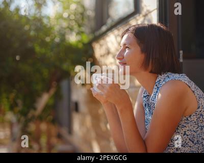 Die Frau trinkt Kaffee, Morgenroutine in der Nähe ihres winzigen Hauses. Erstes Grundstück. Kleines Apartment im Sommergarten. Minimalismus. Wir ziehen ein. Allein zu leben. Charmantes Wohnwagenhaus Stockfoto