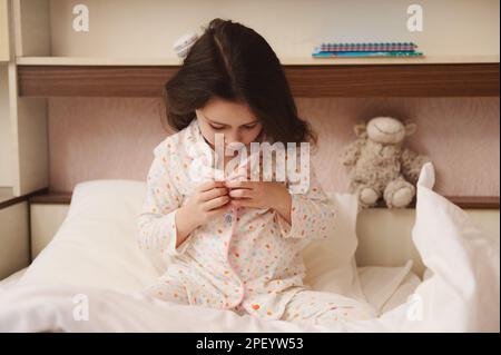 Ein charmantes kleines Mädchen macht ihren Pyjama zu, setzt sich ins Bett und macht sich fürs Bett fertig. Das Konzept, ins Bett zu gehen und aufzuwachen Stockfoto