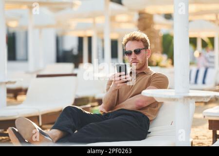 Sitzt und benutzt das Telefon. Der junge Mann ist tagsüber draußen. Urlaubskonzept Stockfoto