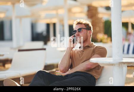 Sitzt und benutzt das Telefon. Der junge Mann ist tagsüber draußen. Urlaubskonzept Stockfoto