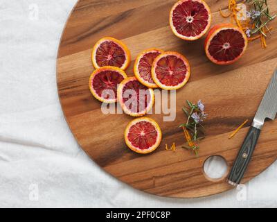 Scheiben von sizilianischem Moro-Blut Orange und Zweige von blühendem Rosmarin auf einem Holzschneidbrett auf einer weißen Tischdecke, die von oben aufgenommen wurde, mit Platz zum Kopieren Stockfoto