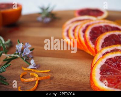 Nahaufnahme von frisch geschnittenen Blutorangen (sizilianische moro) auf einem Schneidebrett mit Zweigen von blühendem Rosmarin und Locken aus Orangenschale Stockfoto