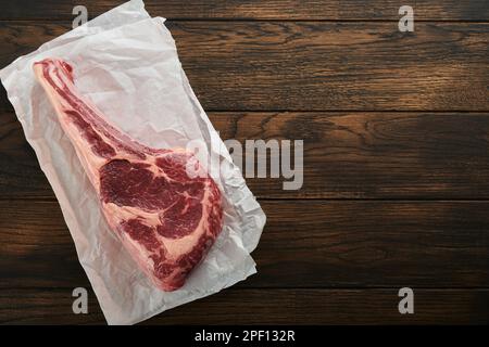 Frisches rohes Tomahawk-Steak. Trocken gereiftes rohes Tomahawk-Rindersteak mit Kräutern und Salz auf altem Holzhintergrund. Grillvorgang wird vorbereitet. Draufsicht und Kopierbereich. Stockfoto