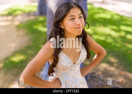 Asiatisches Mädchen im Teenageralter mit Hüften und einem dickköpfigen Look | Ballkleid | Stehend unter einem bemalten blauen Baum auf einem Platz Stockfoto