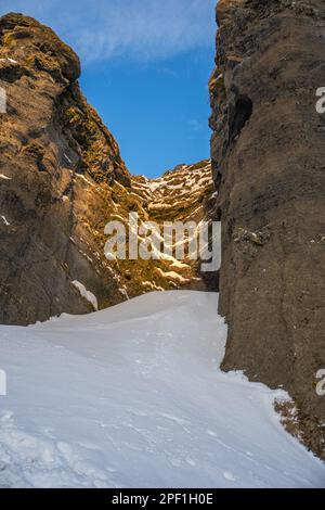 Total schneebedeckte Straße zwischen den Bergen in Richtung des Kvernufoss Wasserfalls und einer Schlucht mit verschneiten Bereichen und mit Sonnenaufgang beleuchtet Stockfoto