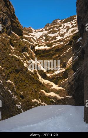 Total schneebedeckte Straße zwischen den Bergen in Richtung des Kvernufoss Wasserfalls und einer Schlucht mit verschneiten Bereichen und mit Sonnenaufgang beleuchtet Stockfoto