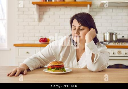 Übergewichtige junge Frau, die zögert, einen riesigen Burger zu essen Stockfoto
