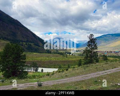 Die Straße entlang des Flusses führt in die Berge von den Hütten des Dorfes Chibit in Altai, Sibirien unter dicken Wolken. Stockfoto