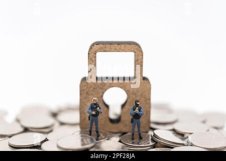 Geld- und Sicherheitskonzept. Zwei Soldat-/Polizeiwächter, Miniaturfiguren, die auf einem Haufen Münzen stehen, mit einem Schlüsselsymbol aus Holz auf weißem Bac Stockfoto