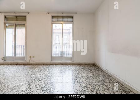 Leeres Wohnzimmer eines Hauses mit Terrazzo-Böden mit schwarzen Punkten, zwei Balkone zur Straße mit Aluminium- und Glasschiebetüren und altem Shuttlebus Stockfoto