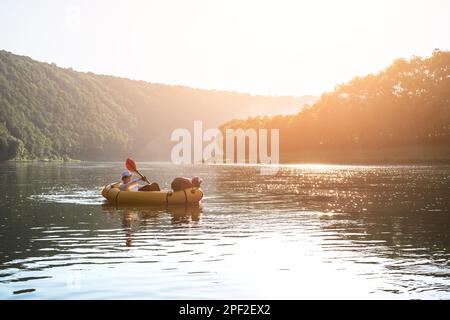 Tourist auf einem gelben Paketboot mit rotem Paddelboot auf einem wunderschönen Fluss bei Sonnenuntergang. Packrafting und Konzept der aktiven Lebenszeit Stockfoto