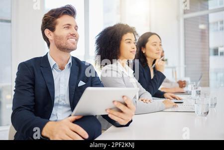 Der junge kaukasische Geschäftsmann, der während einer Präsentation in einem Sitzungssaal mit seinen verschiedenen Kollegen im auf einem digitalen Tablet-Gerät surft Stockfoto