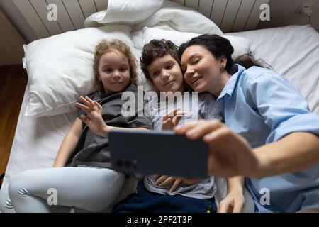 Glückliche weiße Mutter und 2 Kinder haben Online-Videoanrufe mit Vater oder Großmutter Handy, wenn sie zu Hause im Bett liegen. Das Konzept der modernen Kommunikation Stockfoto