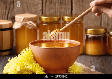 Kreative Zusammensetzung von Gläsern mit Honig, Honig in Tonschüssel, Pendelarm und Honiggießen auf Holzuntergrund. Honig wird aus einem Holzlöffel ausgeschenkt Stockfoto