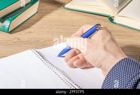 Die Hand eines Mannes mit einem blauen Stift macht Notizen in einem offenen Notizbuch auf einem Büroschreibtisch Stockfoto