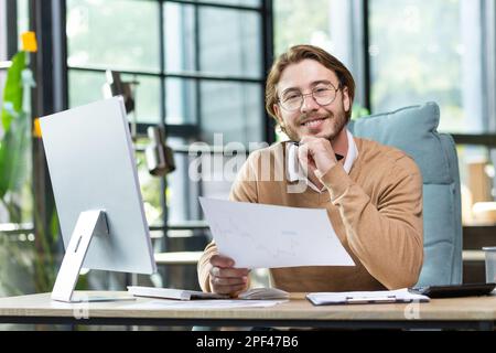 Erfolgreicher und selbstbewusster junger Geschäftsmann, Freiberufler arbeitet in einem modernen Büro mit Dokumenten und Papieren. An einem Schreibtisch mit einem Computer sitzen. Lächelnd in die Kamera. Stockfoto
