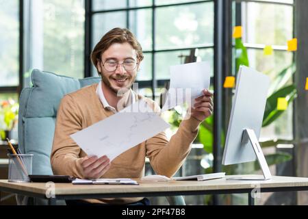 Ein junger Geschäftsmann, ein Freiberufler, arbeitet in einem modernen Büro mit Dokumenten und Papieren. An einem Schreibtisch mit einem Computer sitzen. Stockfoto