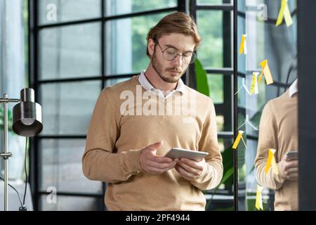 Junger fokussierter männlicher Student, Praktikant im Online-Kurs, der auf den Tablet-Bildschirm schaut, neben einem transparenten Whiteboard mit Notizen steht und sich eine Videokurse ansieht. Stockfoto