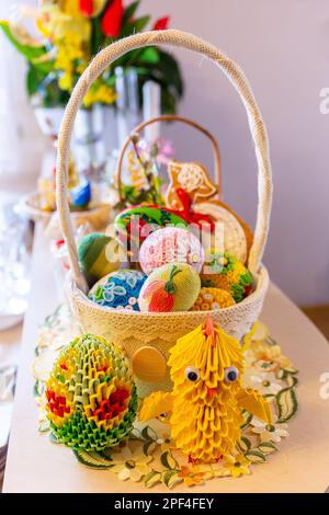 Der Tisch ist mit einem traditionellen Osterkorb mit farbenfrohen, handgefertigten Osteroriginami-Eiern, Küken, Blumen und anderen Osterdekorationen dekoriert. Stockfoto
