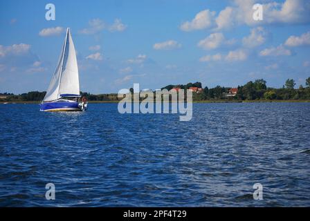 Ein blaues Segelboot segelt auf dem See. Die Küste und einige Häuser im Hintergrund. Yachtfahrt in Masurien (Mazury). Masurian Lake District, Polen. Stockfoto