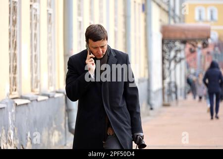 Verärgerter Mann in einem Mantel, der auf dem Smartphone sprach, während er die Straße entlang ging. Handy-Nutzung in der Frühlingsstadt Stockfoto