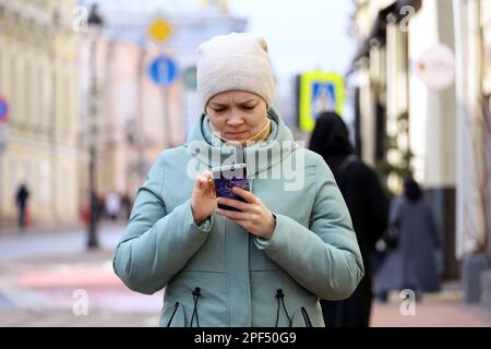 Frau mit Mantel und Hut, die auf einer Straße Smartphone liest. Handy-Nutzung in der Frühlingsstadt Stockfoto