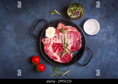 Rohe saftige Steaks mit Gewürzen in einer schwarzen Pfanne, bereit zum Rösten auf rustikalem Betonhintergrund. Frische marmorierte Fleischsteaks mit Kräutern, Knoblauch, Oliven Stockfoto