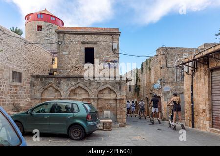 Rhodos, Griechenland - 24. August 2022: Blick auf die Altstadt von Rhodos, Griechenland. Gepflasterte Straßen und Gehwege mit Steinhäusern. Eine Gruppe von Personen macht eine Stadtbesichtigung auf einem dreirädrigen Motorroller. Stockfoto