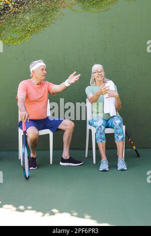 Fröhliche weiße Seniorenfreunde, die auf Stühlen sitzen und sich am sonnigen Tag auf dem Tennisplatz unterhalten, Kopierraum Stockfoto