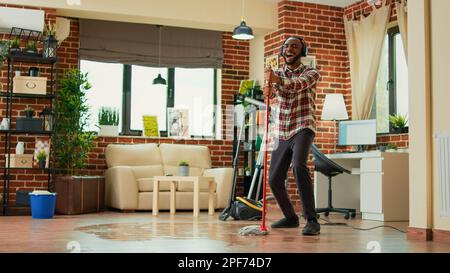Junger männlicher Erwachsener, der zu Hause Spaß mit Mopp und Musik hat, Tanzbewegungen zeigt und im Wohnzimmer singt. Glückliche Person, die Holzböden gewischt und Frühjahrsputz und Hausarbeit macht. Stockfoto