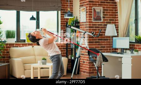 Kaukasische Frau, die sich albern benimmt, Holzböden wischt, Schmutz und Staub mit Mopp fegt und Spaß hat. Junge Person, die zu Hause Musik hört und singt, die Wohnung putzt. Stativaufnahme. Stockfoto