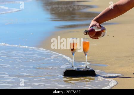 Einschenken von Rosenchampagner oder Cava Sekt, serviert auf Stein am weißen Sandstrand mit tropischem Wasser und blauem Meer, romantischer Urlaub, Wintersonne an Stockfoto