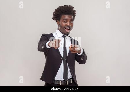 Schockierter Mann mit Afro-Haarschnitt, der mit dem Zeigefinger auf die Kamera zeigt, mit großer Überraschung, Atemanhalten zeigt nach vorne, trägt weißes Hemd und Smoking. Studioaufnahmen im Innenbereich isoliert auf grauem Hintergrund. Stockfoto