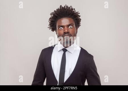 Porträt eines Mannes mit Afro-Haarschnitt mit wütendem, strengen Gesichtsausdruck, der direkt in die Kamera schaut, negative Emotionen hat, weißes Hemd und Smoking trägt. Studioaufnahmen im Innenbereich isoliert auf grauem Hintergrund. Stockfoto