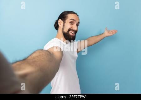 Porträt eines königsfreundlichen Mannes mit Bart, der ein weißes T-Shirt trägt, mit einem Lächeln in die Kamera schaut, eine Willkommensgeste macht, Selfie-POV macht. Innenstudio, isoliert auf blauem Hintergrund. Stockfoto