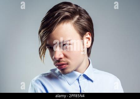 Porträt eines jungen, attraktiven Mannes mit langen Fransen auf den Augen, der mit mysteriösem Blick wegsieht, ein blaues Hemd trägt. Studioaufnahmen im Innenbereich isoliert auf grauem Hintergrund. Stockfoto