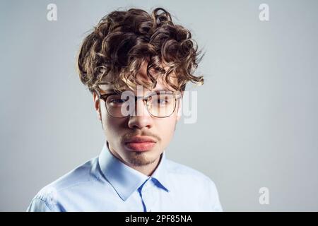 Porträt eines ernsthaften jungen Erwachsenen mit blauem Hemd und Brille, lockiger Frisur, der mit striktem Gesichtsausdruck in die Kamera schaut. Studioaufnahmen im Innenbereich isoliert auf grauem Hintergrund. Stockfoto