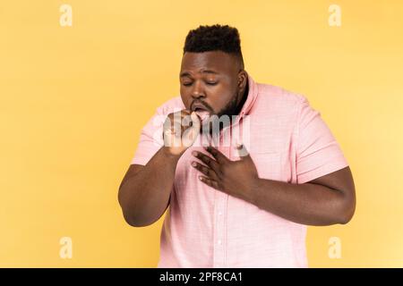 Porträt eines kranken, ungesunden, bärtigen Mannes, der ein rosa Hemd trägt, Husten und Schmerzen in der Lunge hat, sich Erkältung holt, Grippesymptome. Innenstudio-Aufnahme isoliert auf gelbem Hintergrund. Stockfoto