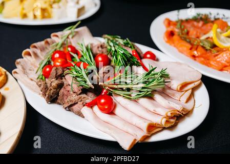 Teller mit dünn geschnittenem Schinken und Roastbeef mit rotem, heißem Paprika und Kirschtomaten und Rosmarin-Ästen. Die Vorspeisenplatten bei der Veranstaltung. Catering Stockfoto