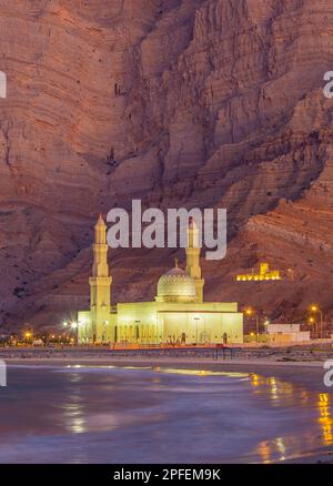 Dramatische felsige Kulisse hinter der beleuchteten Khasab-Moschee zur Blue Hour in Khasab, Oman Stockfoto