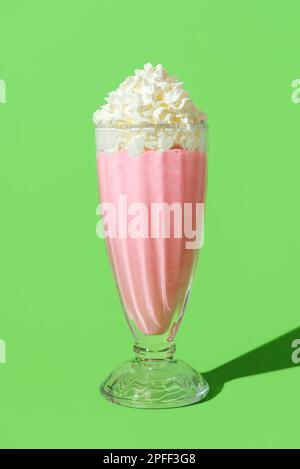 Nahaufnahme mit einem Glas Erdbeermilchshake garniert mit Schlagsahne. Köstlicher Milchshake in hellem Licht minimalistisch auf einem Tisch in leuchtendem Grün. Stockfoto