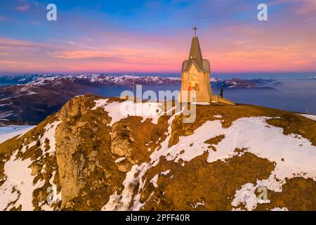 Der Winteruntergang über der Kapelle Monumento al Redentore auf dem Gipfel des Mount Guglielmo aus der Vogelperspektive. Zone, Provinz Brescia, Lombardei, Italien. Stockfoto