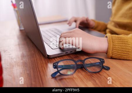 Nahaufnahme der Hände eines Kindes auf einem Laptop, wobei der Finger den Cursor auf dem Trackpad bewegt. Der Holzschreibtisch im Hintergrund hat ein Paar FO Stockfoto