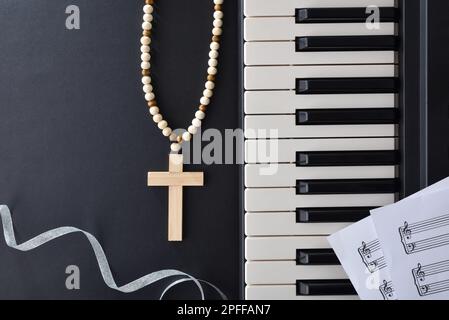 Hintergrund für christliche Musik mit Klaviermusik und Notenblättern auf einem schwarzen Tisch mit Kreuz und silberner Schleife. Draufsicht. Stockfoto